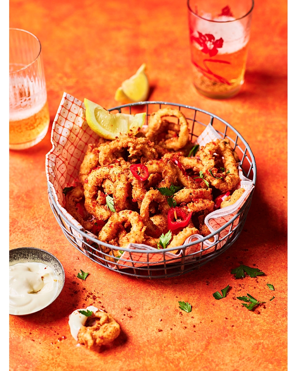 Overhead shot of fried calamari in a metal basket, with lemon wedges, dipping sauce, parsley, and beer on an orange surface.