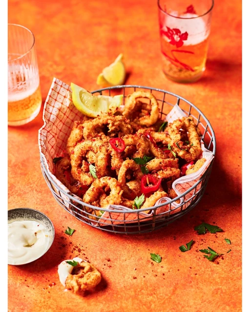 Overhead shot of fried calamari in a metal basket, with lemon wedges, dipping sauce, parsley, and beer on an orange surface.