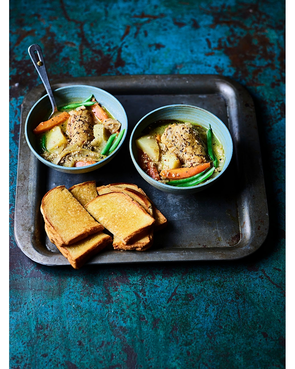 Overhead shot of two bowls of chicken stew with vegetables and slices of toast on a vintage metal tray set against a teal background.