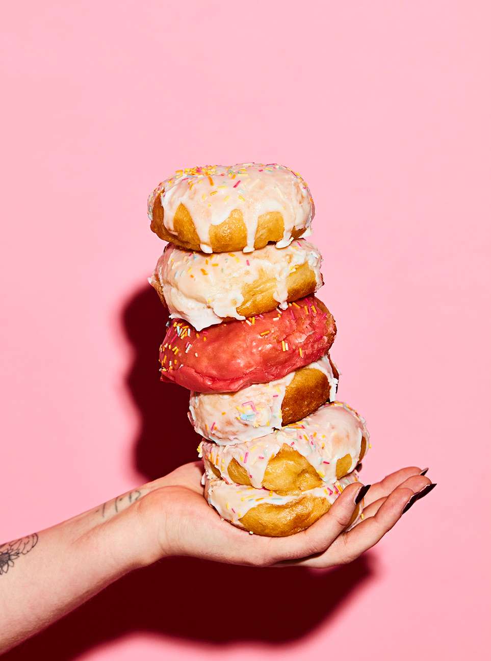 Stacked donuts held by a hand against a pink backdrop, featuring colorful sprinkles and icing with a playful, confectionery theme.