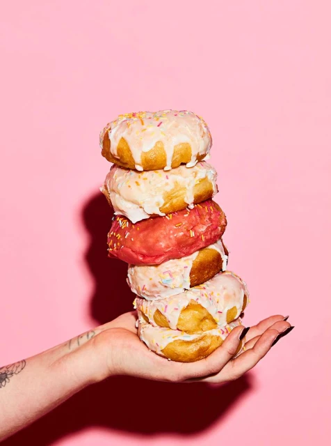 Stacked donuts held by a hand against a pink backdrop, featuring colorful sprinkles and icing with a playful, confectionery theme.