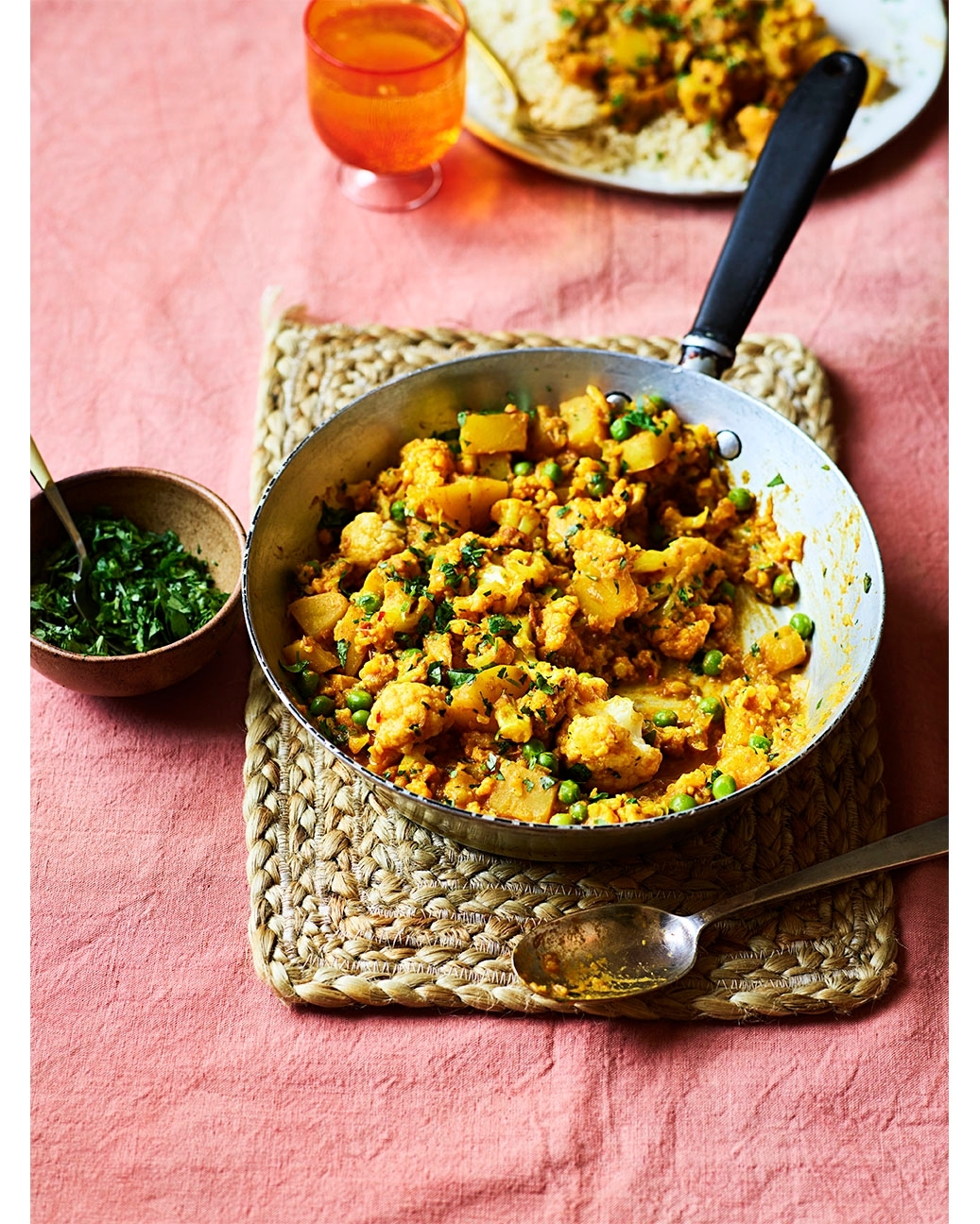 Overhead shot of a frying pan with cauliflower and potato curry on a jute placemat, set on a pink tablecloth, with a glass of orange juice.