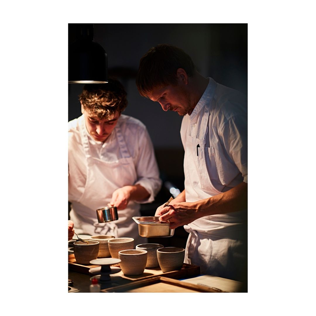 Two chefs in white uniforms preparing food in a dimly lit kitchen, with various small bowls and cooking utensils on the counter.