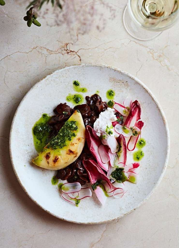 Overhead shot of a rustic white plate with grilled melon, ricotta, radicchio, and a green sauce drizzle, on a textured beige surface.