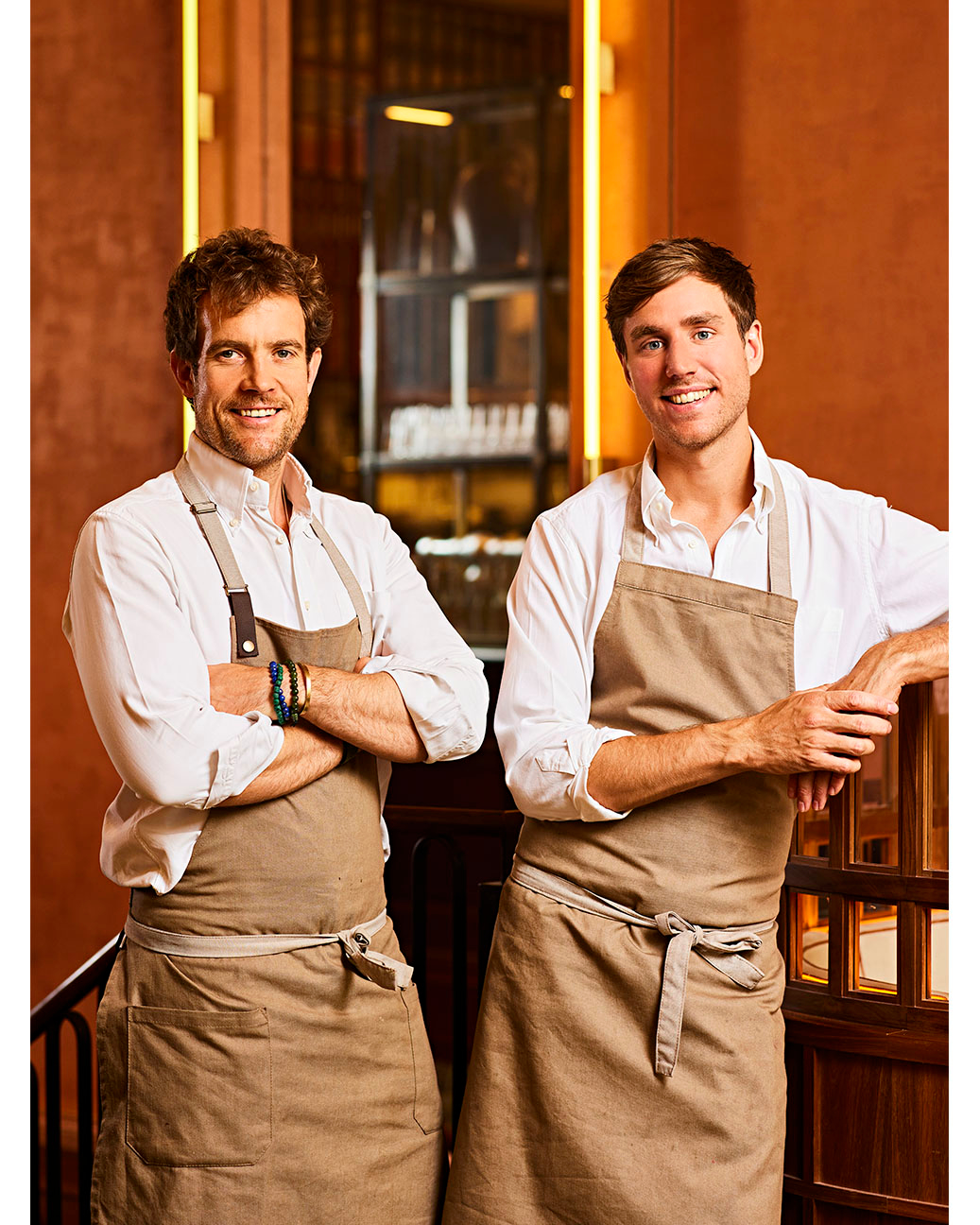 Two chefs in aprons pose indoors in warm lighting. A vertical shot features two smiling men in chef's aprons, standing in front of shelving.