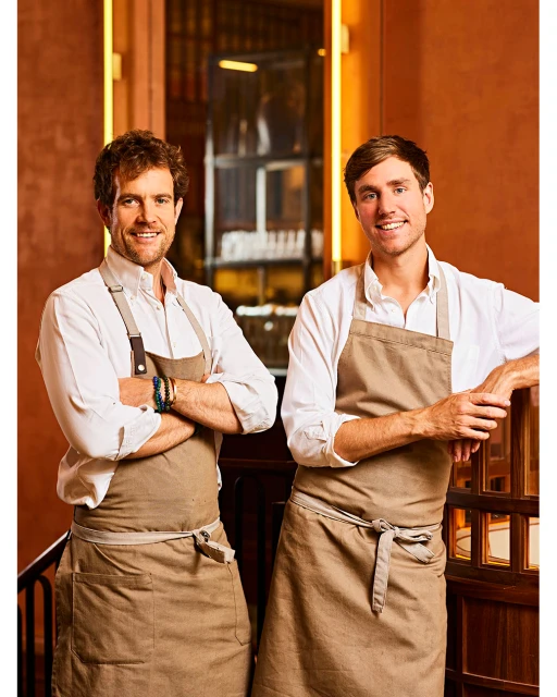 Two chefs in aprons pose indoors in warm lighting. A vertical shot features two smiling men in chef's aprons, standing in front of shelving.