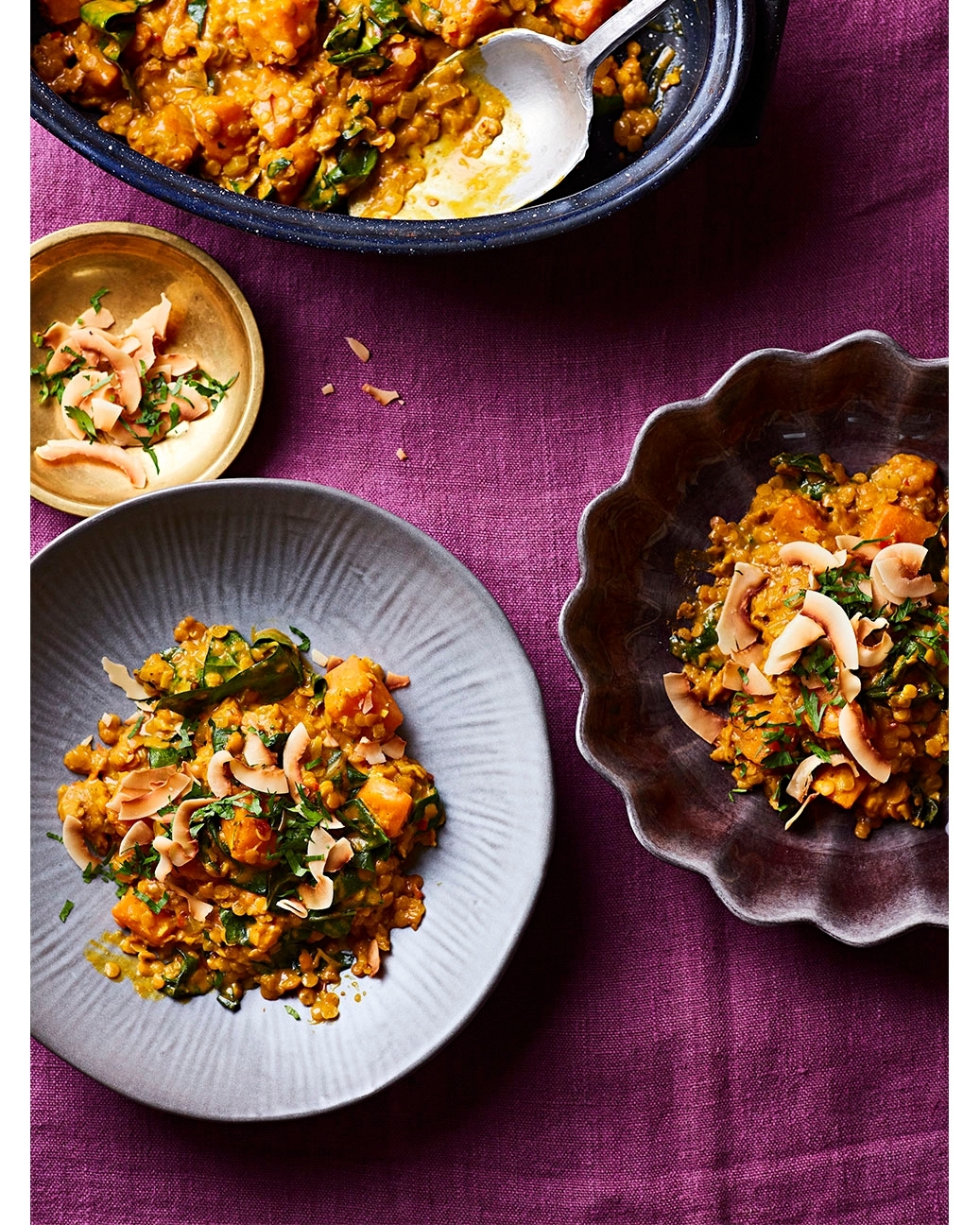 Overhead shot of two plates of lentil and butternut squash curry with coconut flakes and chopped greens on a magenta-colored tablecloth.
