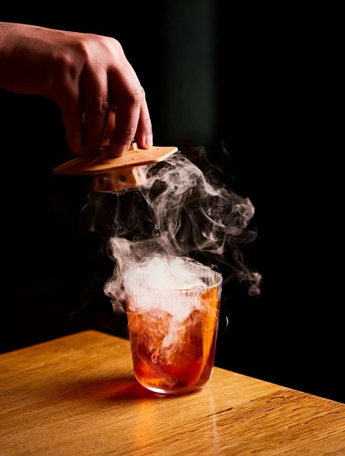 Close-up of a cocktail being served with smoke in a transparent glass on a wooden table with a black background. 