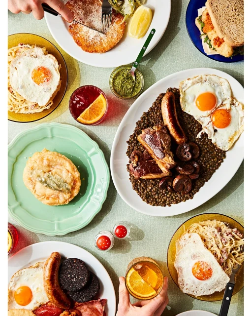 Overhead shot of multiple plates filled with various brunch foods, including eggs, sausages, lentil stew, and bread, arranged on a table.