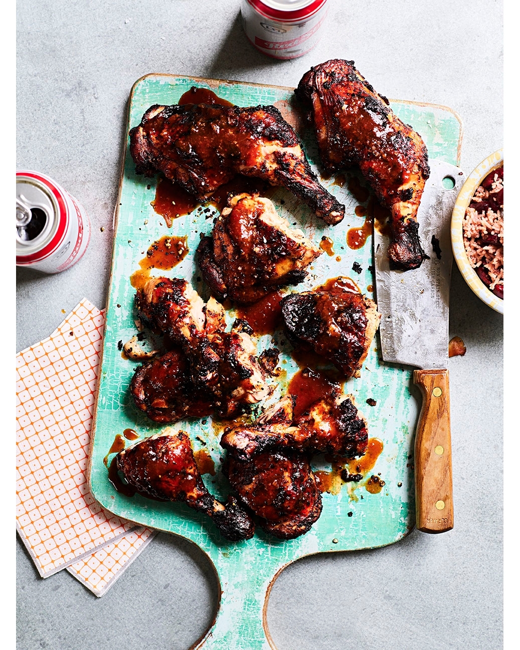 Overhead shot of glazed chicken drumsticks arranged on a rustic turquoise cutting board with a bowl of beans and soda cans.