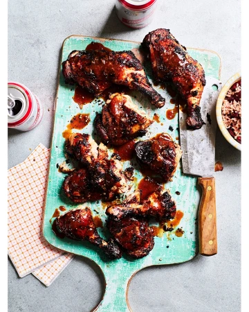 Overhead shot of glazed chicken drumsticks arranged on a rustic turquoise cutting board with a bowl of beans and soda cans.