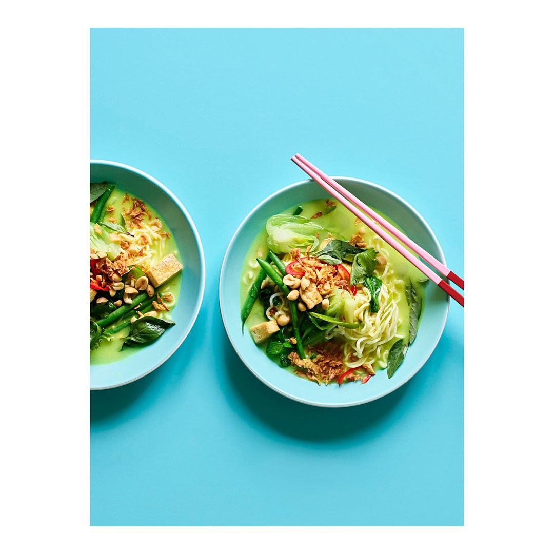 Overhead shot of two bowls of noodle soup with vegetables and pink chopsticks on a bright blue background, flat lay food photography.