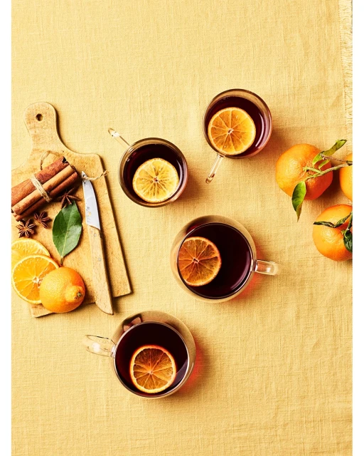 Overhead shot of four glasses of mulled wine garnished with orange slices, arranged on a yellow tablecloth with cinnamon sticks and oranges.