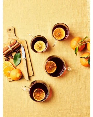 Overhead shot of four glasses of mulled wine garnished with orange slices, arranged on a yellow tablecloth with cinnamon sticks and oranges.