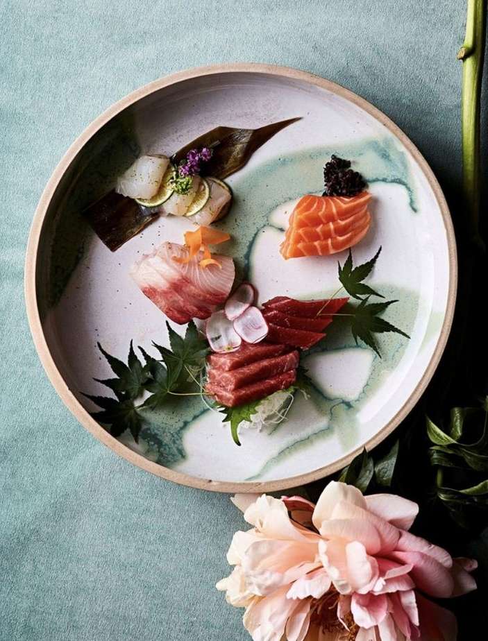 Overhead shot of a round plate of assorted sushi and sashimi, garnished with flowers and leaves, next to a peach peony.