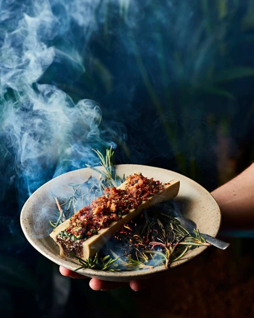 Overhead shot of seasoned bone marrow on a plate held by a hand, garnished with rosemary, and encircled by smoke for a unique presentation.