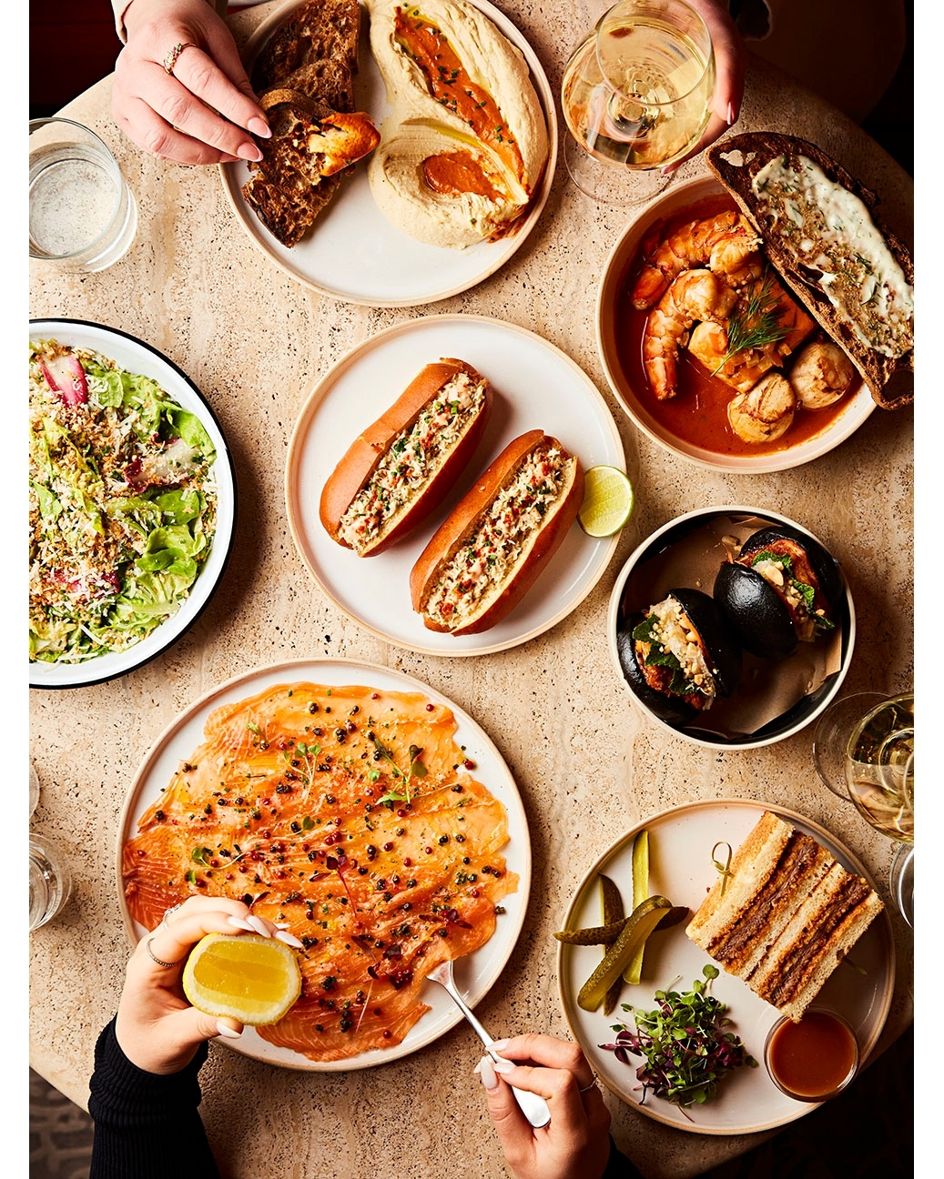 Overhead shot of a variety of prepared food on plates and hands holding a fork and lemon wedge, ready to serve and eat.
