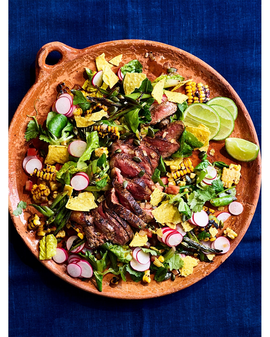 Overhead shot of a vibrant steak salad on a rustic clay plate with lime wedges, set against a dark blue textile backdrop.