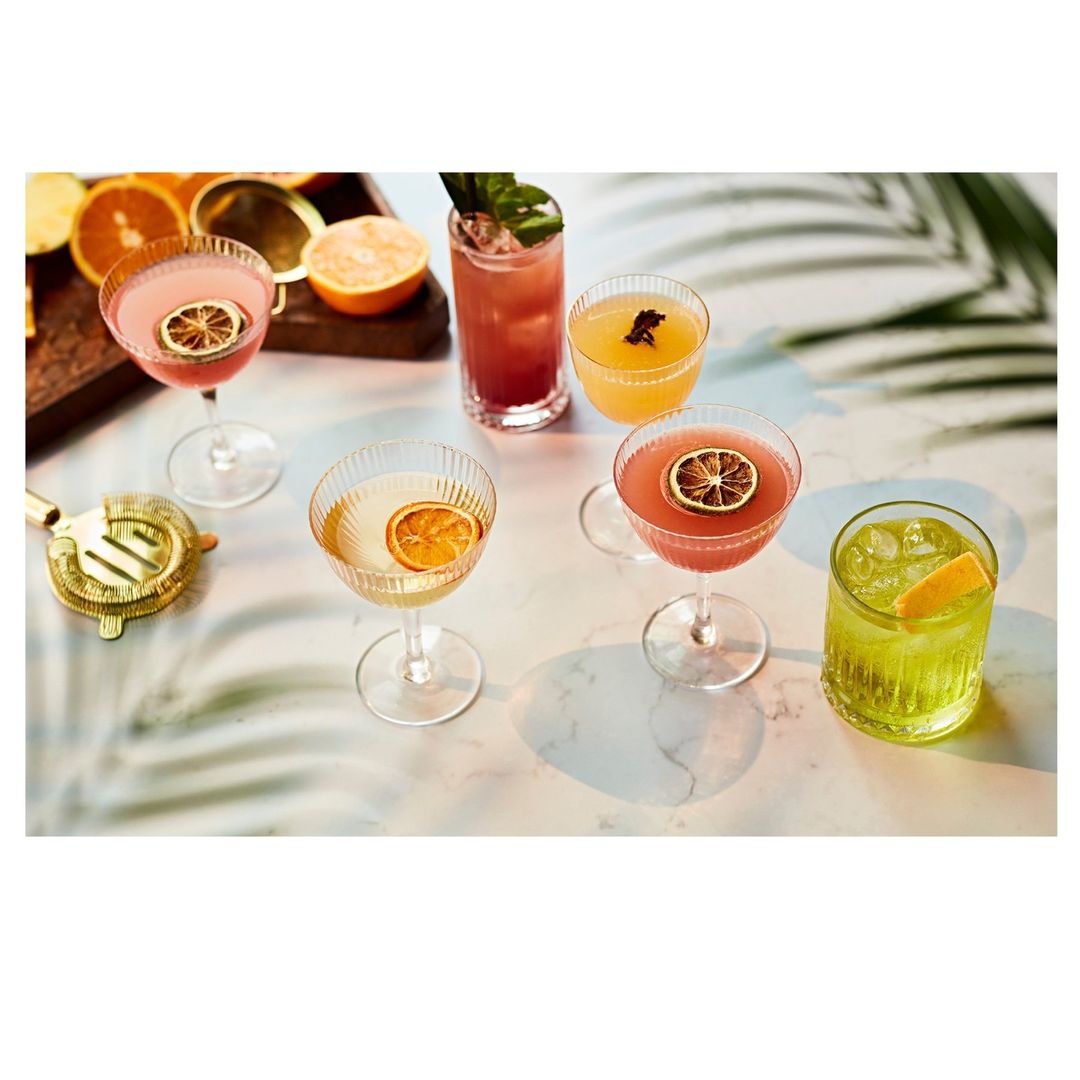 Several colorful cocktails are arranged on a table, garnished with citrus slices, alongside fresh oranges and a gold cocktail utensil.