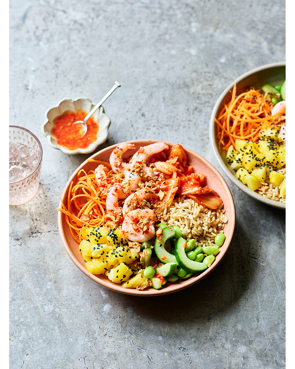Two bowls of shrimp poke with rice, pineapple, edamame, and shredded carrots. One bowl is in focus, the other out of focus.