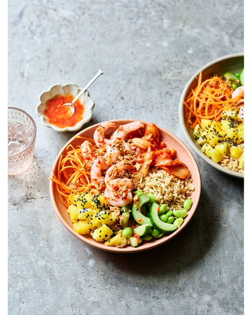 Two bowls of shrimp poke with rice, pineapple, edamame, and shredded carrots. One bowl is in focus, the other out of focus.