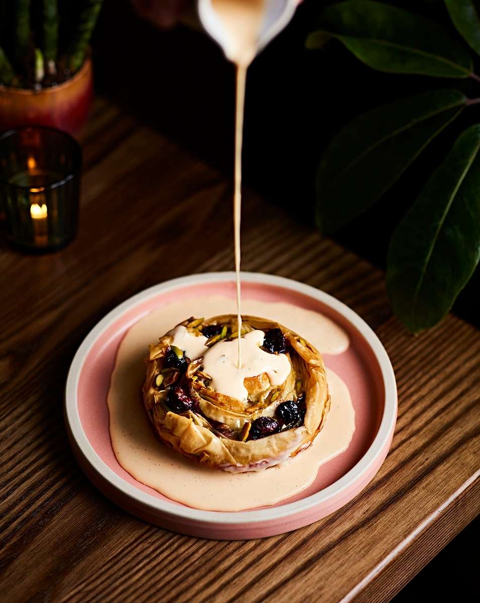 Elevated angle captures sauce being poured over a pastry on a pink plate, displayed on a wooden table, with greenery and candle.