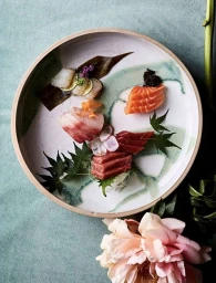 An overhead food photograph of assorted sashimi arranged artistically on a round ceramic plate with green leaves and a pink flower in the foreground.
