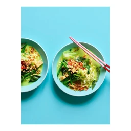 Two blue bowls of noodle soup with green vegetables and pink chopsticks on a bright turquoise background