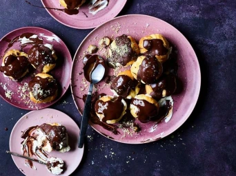 Overhead food photography of chocolate-glazed profiteroles served on pink plates over a dark textured surface.