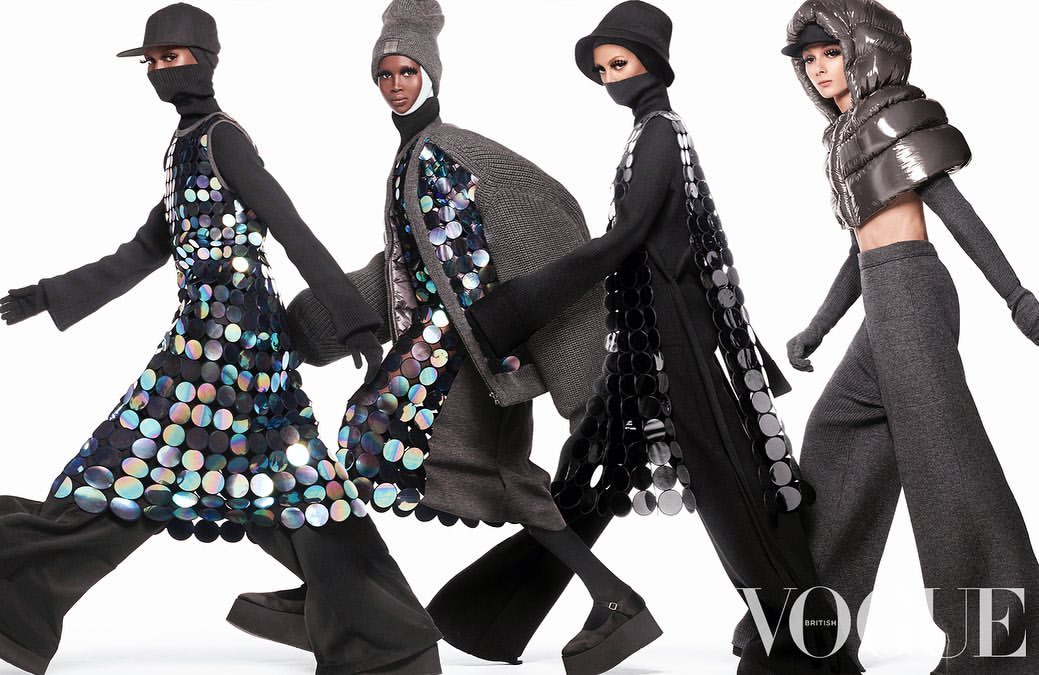 Four fashion models wearing hats and futuristic outfits with reflective discs pose in front of a white studio backdrop, Vogue logo visible.