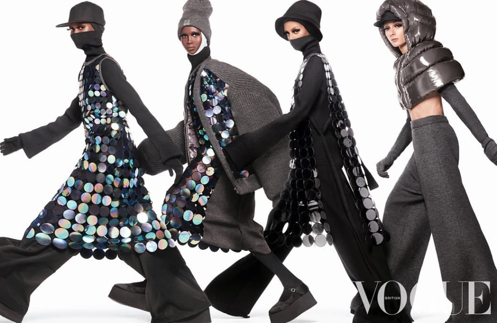 Four fashion models wearing hats and futuristic outfits with reflective discs pose in front of a white studio backdrop, Vogue logo visible.