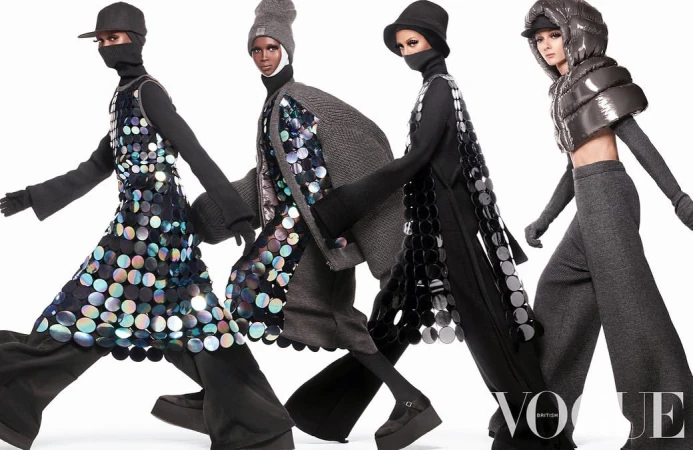 Four fashion models wearing hats and futuristic outfits with reflective discs pose in front of a white studio backdrop, Vogue logo visible.