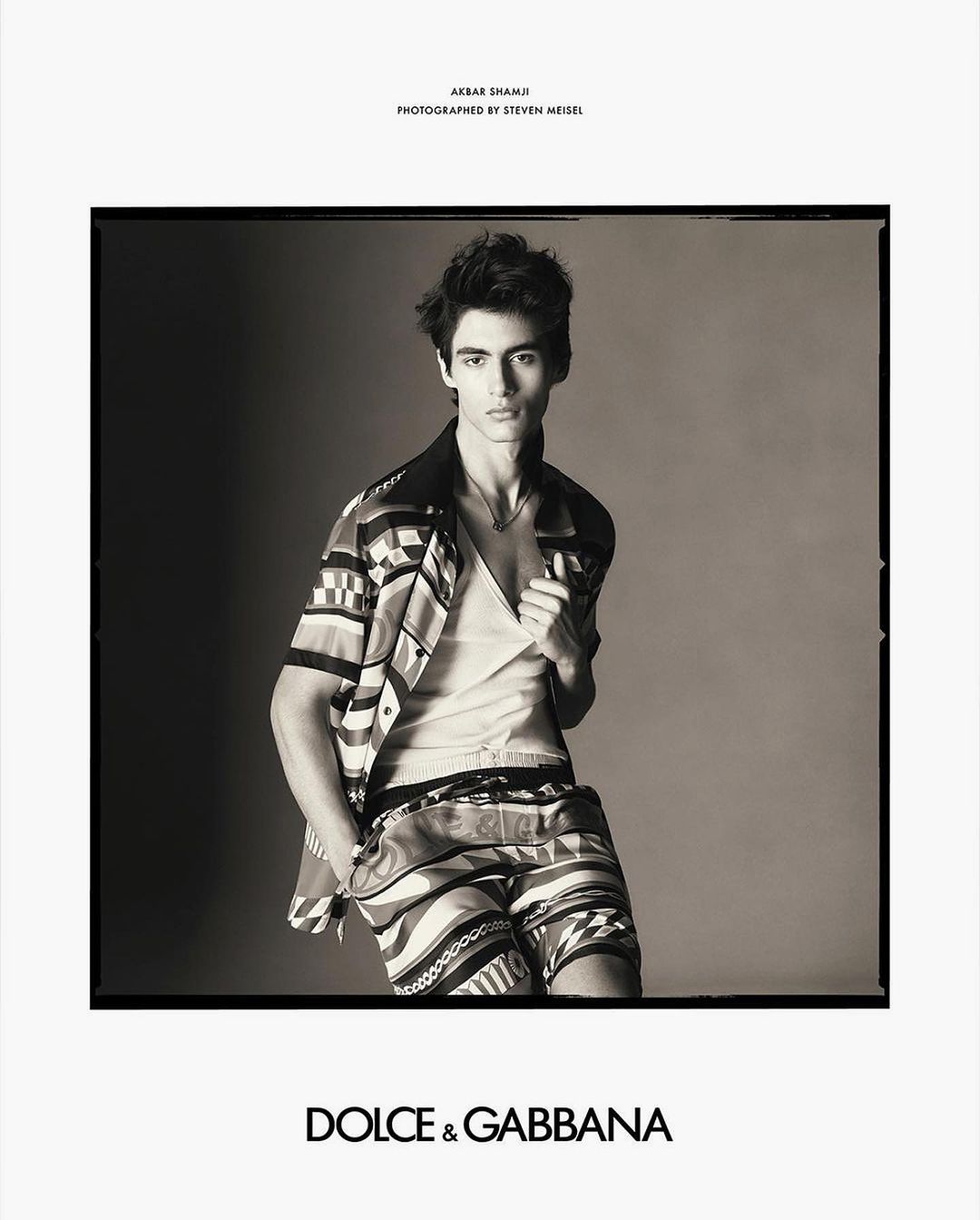 Black and white studio portrait of a young man in a patterned shirt and shorts, looking at the camera, Dolce & Gabbana logo visible.