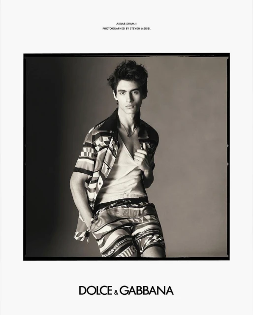Black and white studio portrait of a young man in a patterned shirt and shorts, looking at the camera, Dolce & Gabbana logo visible.