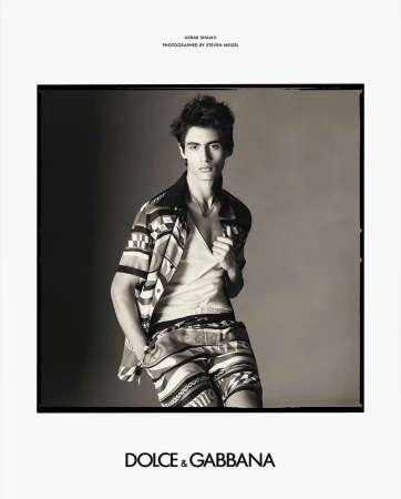 Black and white studio portrait of a young man in a patterned shirt and shorts, looking at the camera, Dolce & Gabbana logo visible.