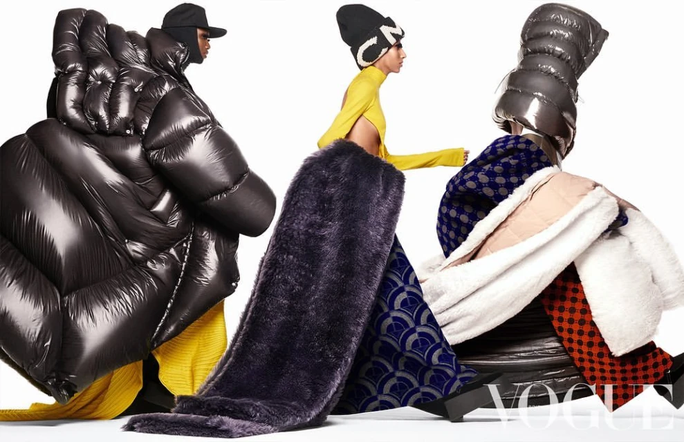Three models showcase avant-garde fashion in a studio shot, featuring oversized puffer coats, unique headwear, and patterned clothing on a white backdrop.