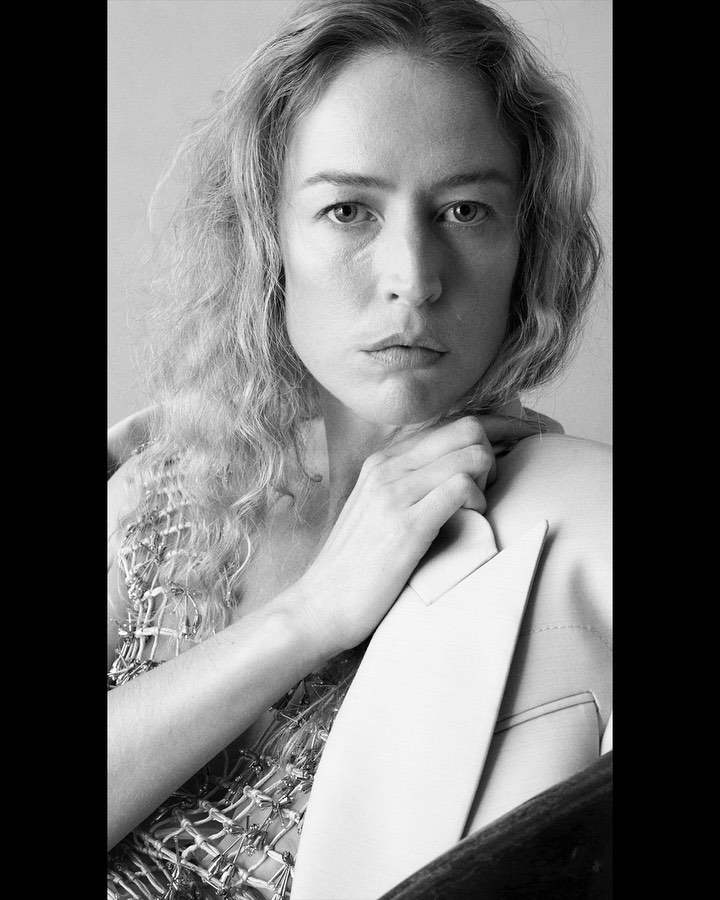 Black and white portrait of a woman with wavy blonde hair, gazing directly at the camera, wearing a textured top and jacket.
