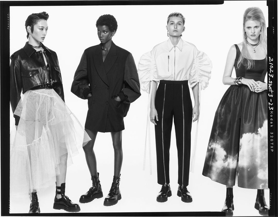 Four fashion models showcasing black and white outfits in a studio shot, featuring tulle skirts, blazers, and accessories against a clean backdrop.