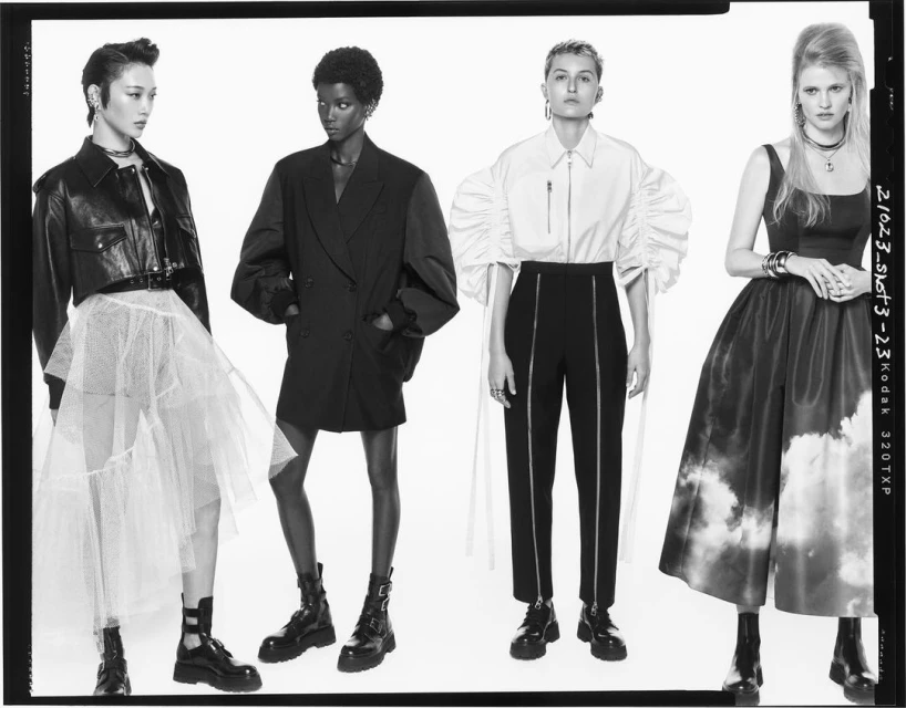 Four fashion models showcasing black and white outfits in a studio shot, featuring tulle skirts, blazers, and accessories against a clean backdrop.