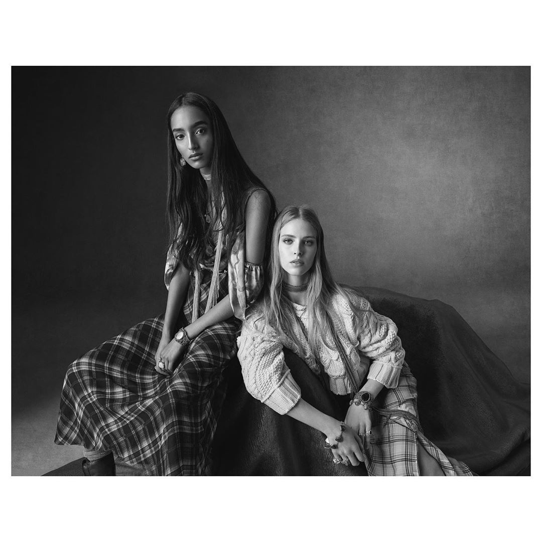 Two fashion models pose together in a studio, wearing patterned dresses, accessorized with watches. The image is monochrome.
