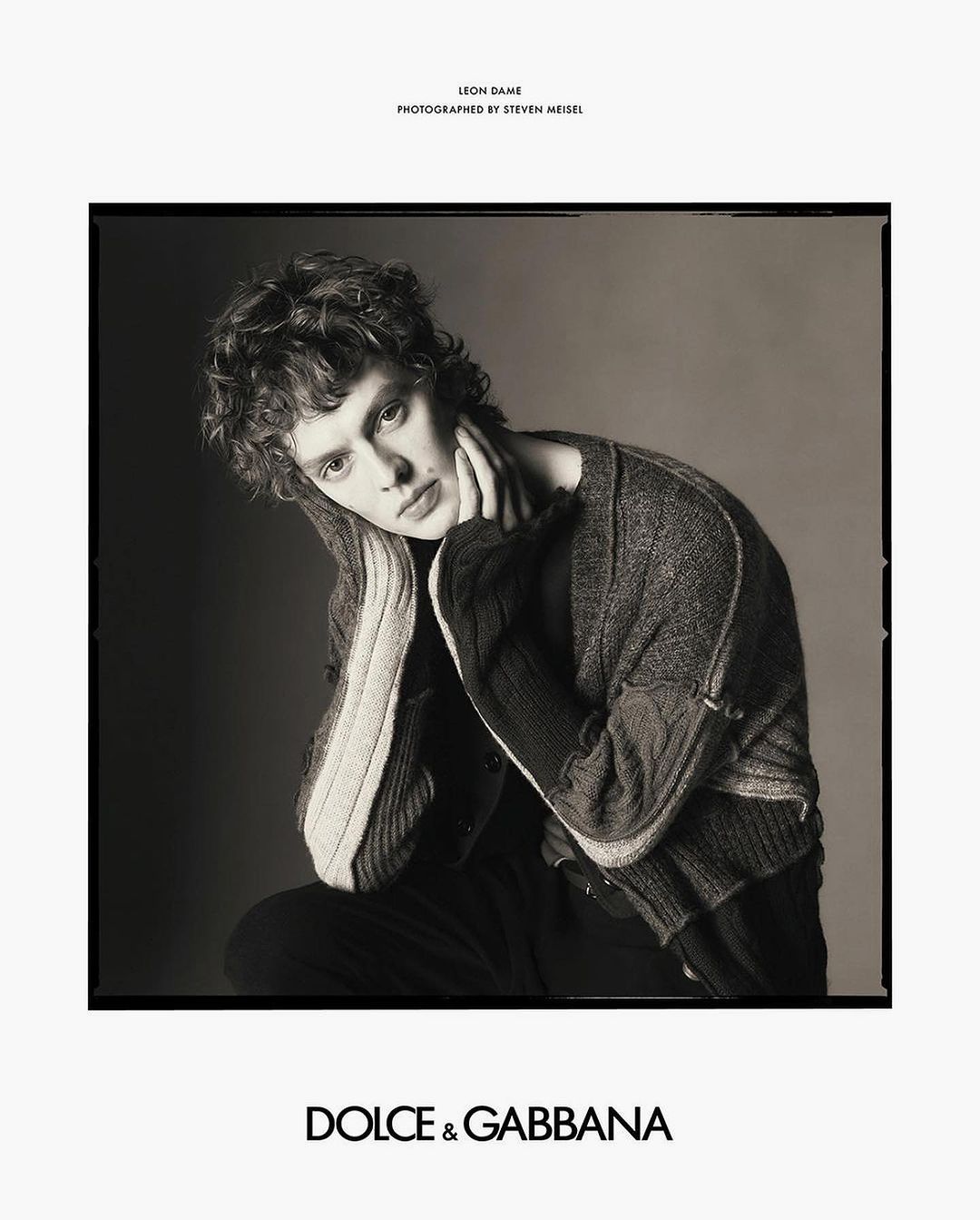 Monochrome fashion portrait of a young male model with curly hair, wearing a cardigan sweater, posing indoors against a plain background.