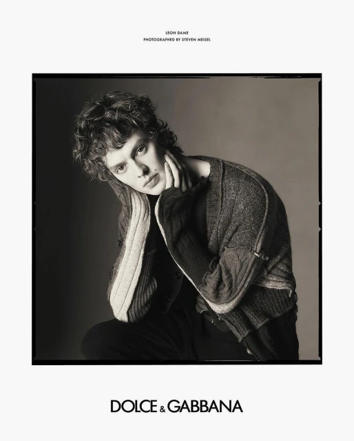 Monochrome fashion portrait of a young male model with curly hair, wearing a cardigan sweater, posing indoors against a plain background.