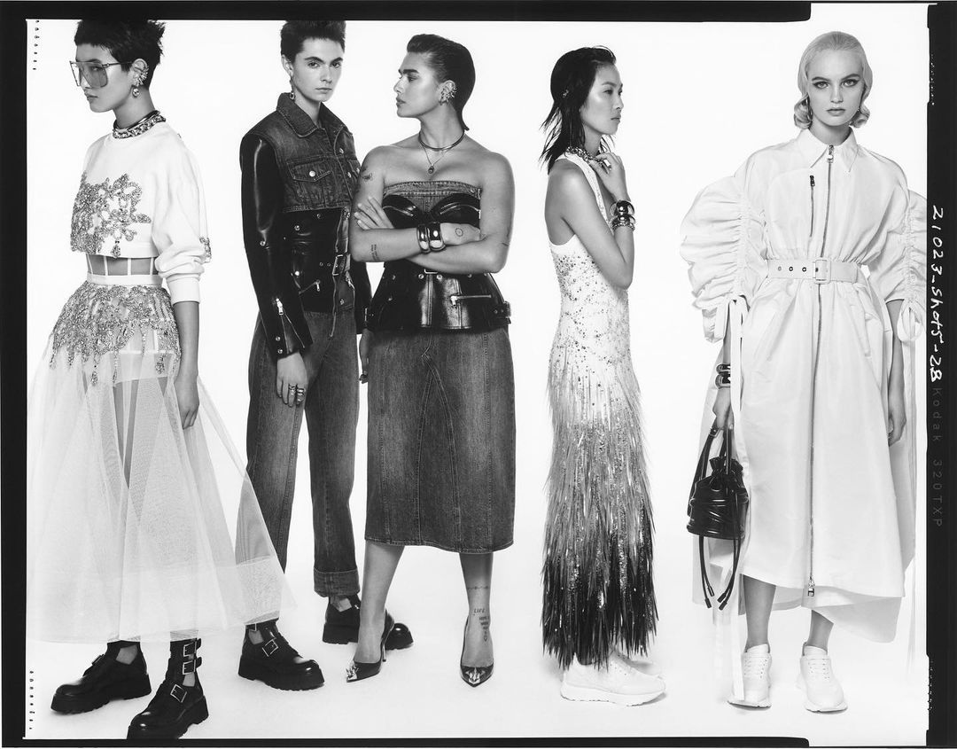 Black-and-white studio shot of five fashion models posing in various designer outfits in a straight line against a stark white backdrop.