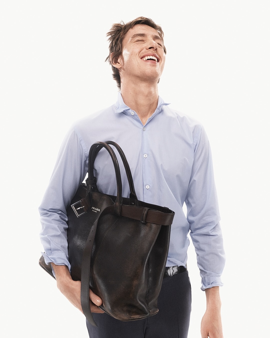 Man smiles in a light blue shirt, carrying a dark brown Prada leather tote bag against a bright white background in a studio shot.