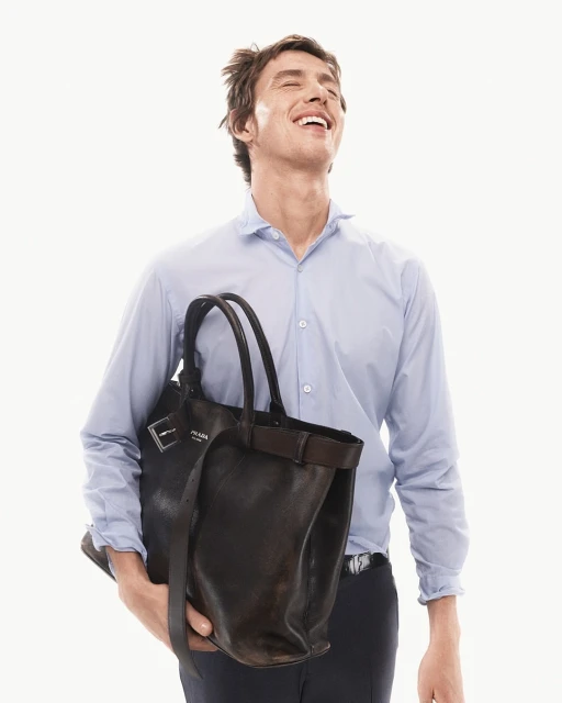Man smiles in a light blue shirt, carrying a dark brown Prada leather tote bag against a bright white background in a studio shot.