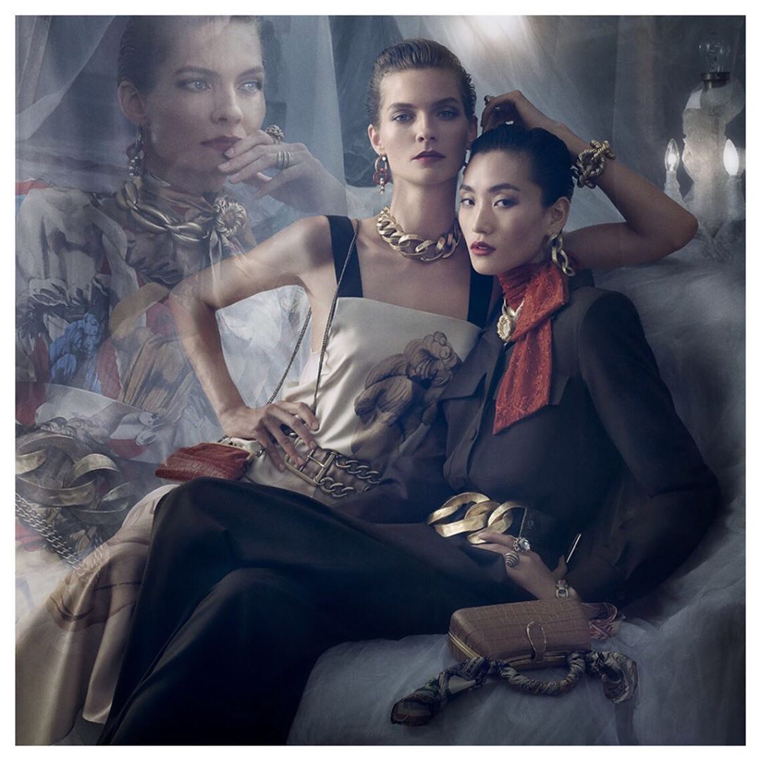 Fashion portrait of two women wearing gold jewelry and dark clothing with a translucent overlaid face in the background by Steven Meisel