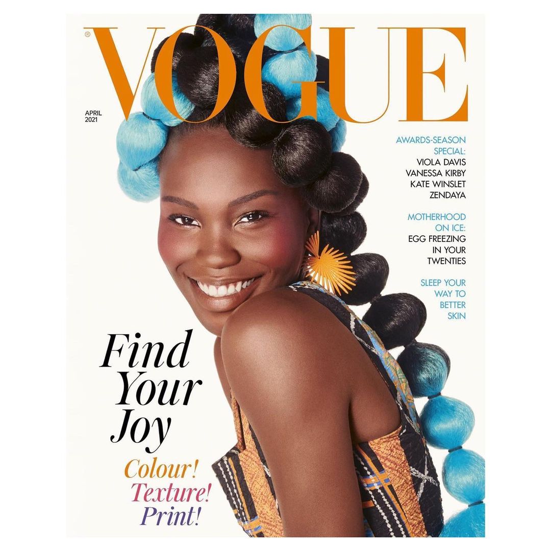 Vogue cover featuring a smiling black woman with elaborate blue and black hair, adorned with an orange earring, set against a white background.