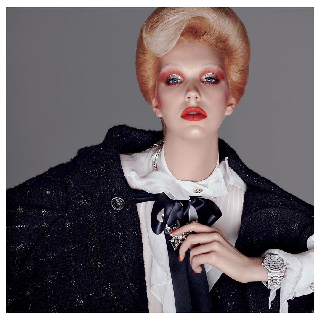 Fashion editorial shot of a blonde model wearing a black tweed jacket, a white ruffled shirt, and an elegant silver watch, posing indoors.