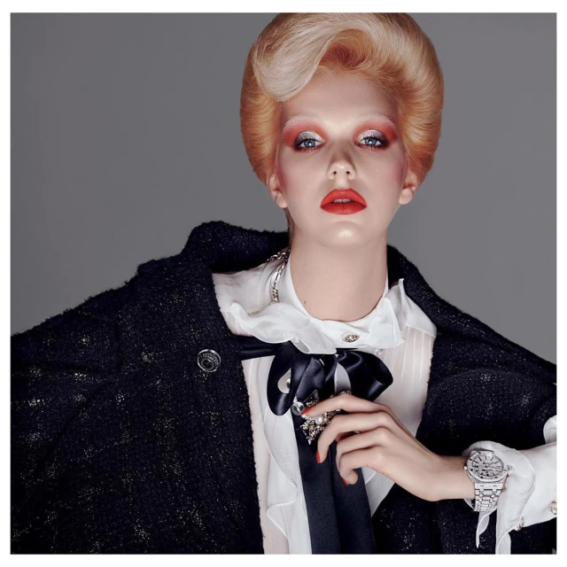 Fashion editorial shot of a blonde model wearing a black tweed jacket, a white ruffled shirt, and an elegant silver watch, posing indoors.