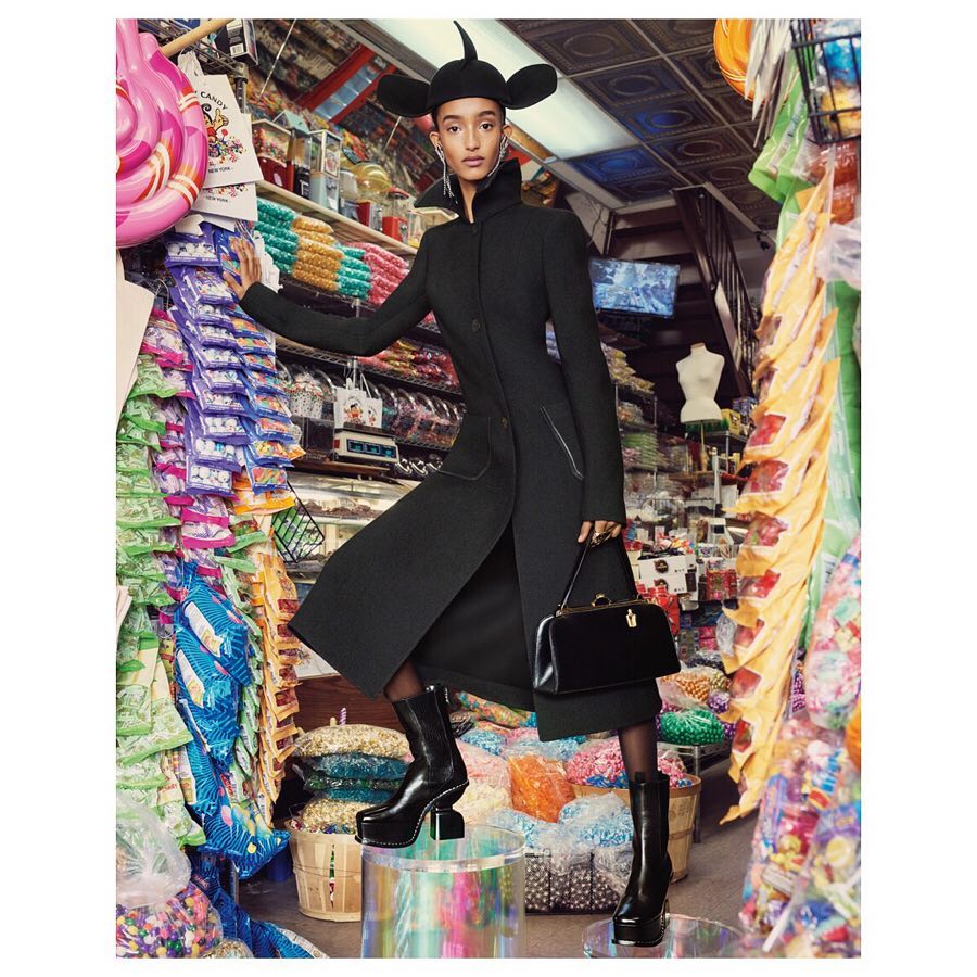 Fashion-forward woman strikes a pose in a unique black ensemble and cartoonish hat inside a candy store, surrounded by colorful sweets.